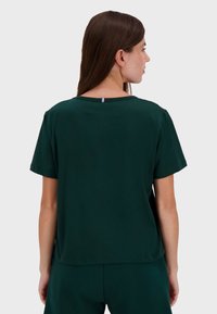 Dark green t-shirt with short sleeves, round neckline, and a small red, white, and blue accent at the back neck. Smooth fabric texture.