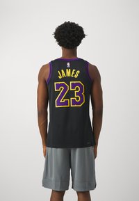 Hombre vistiendo una camiseta de baloncesto negra y morada con "JAMES 23" en la espalda y pantalones cortos grises, de pie contra un fondo blanco liso.