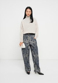 Svetlo béžový rolák skombinovaný s vzorovanými šedými širokými nohavicami. Modelka drží bielu kabelku. Čierne vysoké topánky dopĺňajú celkový vzhľad.