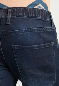 Detailní záběr na kapsu tmavě modrých džínů se švy, nošeno s zasunutým béžovým a modrým topem.