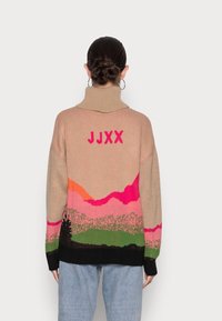 Strickpullover mit beigem Grund, der farbenfrohe Berglandschaftsmuster in Pink, Orange und Grün aufweist. Hoher Kragen, lockere Passform, schwarze Akzente.