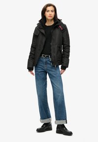 Czarna izolowana kurtka z zapięciem na zamek, ściągaczami przy mankietach i dwiema bocznymi kieszeniami. Połączona z luźnymi niebieskimi dżinsami i czarnymi sznurowanymi butami.