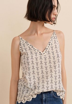 Vrouw met kort donker haar die een mouwloze crème top draagt met een zwart patroon en geschulpte zoom, gecombineerd met blauwe spijkerbroek, zijwaarts gedraaid.
