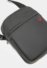 Mustikka, sileäpintainen olkalaukku vetoketjullisella sulkemisella, jossa on etutasku ja kontrastina punainen logo. Yksinkertainen suorakulmainen muotoilu.