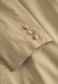 Beige szövet közeli felvétel, hajtott gallérral és három áttetsző gombbal, amelyeken középen nyílás található, sima textúrával és tiszta varrásokkal.
