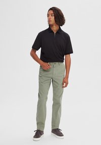 Camisa polo negra con collar de cremallera; combinada con pantalones verde claro. Las zapatillas grises completan el conjunto, resaltando tonos neutros y un estilo casual.