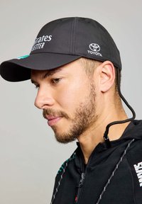 Cappellino sportivo nero con visiera curva, dotato del logo "Emirates" e dell'emblema Toyota. Il cappellino presenta un tessuto testurizzato e un cordino di regolazione sul retro.