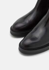 Botas de tobillo de cuero negro con un diseño elegante, punta redondeada y una suela de goma negra contrastante para tracción. Textura suave con un brillo sutil.