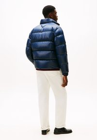 Mand iført en skinnende navy puffjakke, hvide bukser og sorte sko, stående med ryggen til en ensfarvet hvid baggrund.
