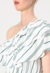 Top alb cu un umăr (one-shoulder) care prezintă modele cu dungi verzi și accente de volane stratificate, evidențiind un material moale, texturat.