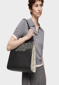 Osoba w szarej koszulce polo z krótkim rękawem i spodniach trzyma dużą czarną torbę z wzorem i złotymi akcentami na ramieniu.