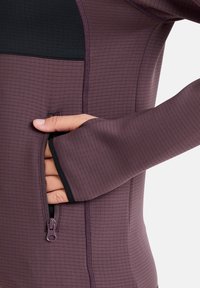 Paarse sportieve top met lange mouwen en een gestructureerd ribbelpatroon, met een ritszak aan de zijkant en zwarte accenten.
