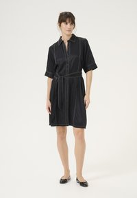 Melnas krekla kleitas ar krekla apkakli, īsām piedurknēm, sašaurinātu jostasvietu un kontrastējošiem baltajiem diegiem. Apvienota ar melniem plakaniem apaviem.