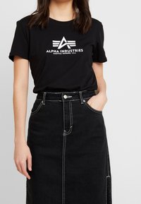 Camiseta de algodón negra con un logo blanco de "Alpha Industries" con alas, combinada con una falda de mezclilla negra de cintura alta con costuras blancas.