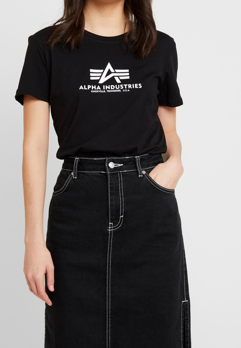 Camiseta de algodón negra con un logo blanco de "Alpha Industries" con alas, combinada con una falda de mezclilla negra de cintura alta con costuras blancas.