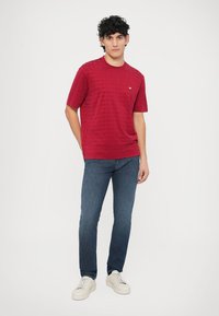 Červené tričko s krátkym rukávom s textúrovaným geometrickým vzorom a malým logom, v kombinácii s modrými džínsami a bielymi teniskami.