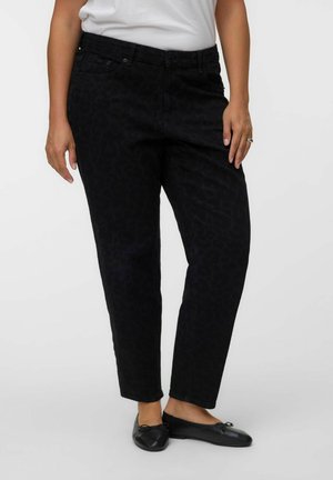 Vrouw draagt zwarte broek met patroon, zwarte ballerina's en een wit topje, staand tegen een effen achtergrond.