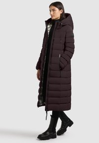 Long burgundy puffer coat with hood, featuring quilted design, silver lining, and zippers, paired with ankle boots.