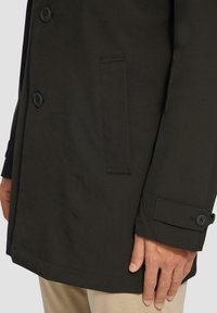 Cappotto nero realizzato in tessuto liscio con design doppiopetto, caratterizzato da grandi bottoni e una tasca laterale. Vestibilità sartoriale.