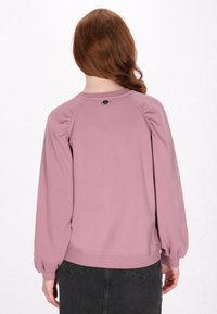 Sweatshirt rosa con maniche a sbuffo, polsini e colletto a costine. Presenta un design arrotondato sul retro con un piccolo dettaglio decorativo nero.