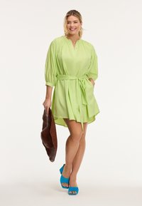 Abito verde chiaro con una vestibilità ampia, maniche corte e una vita con lacci. Completato da tacchi aperti blu e una borsa lavorata a maglia marrone.