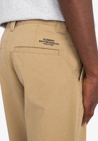 Khaki nohavice vyrobené z bavlneného twillu s hladkým povrchom. Majú zadné vrecká a čierne vyšívané logo na pravom vrecku.