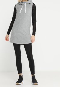 Osoba w szarej sukience z bezrękawnikiem nosi czarną przezroczystą bluzkę z długim rękawem, czarne legginsy oraz czarne buty wsuwane z białymi podeszwami.