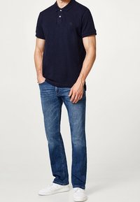 Marinblå polo t-shirt med uppvikt ärm, matchad med blå denimjeans och vita sneakers. Har två knappar och en liten emblem på bröstet.