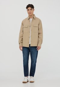 Camicia beige a bottoni con due tasche sul petto, realizzata in tessuto, abbinata a jeans blu scuro e sneakers bianche con suole marroni.