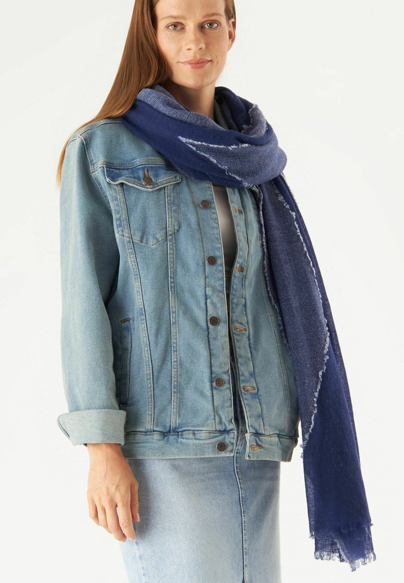 Veste en denim avec fermeture à boutons, couleur bleu délavé, associée à une écharpe texturée bleu marine avec des bords effilochés.