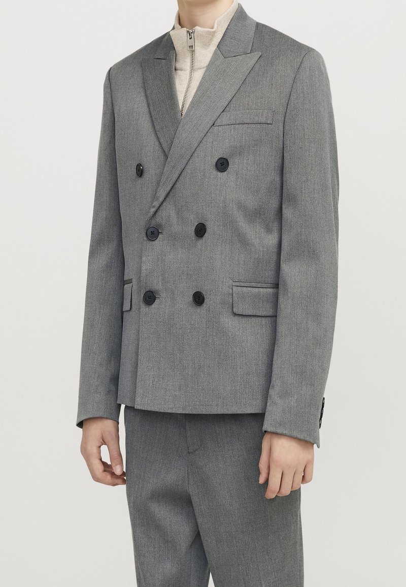 Homme portant une veste de costume croisée grise à boutons noirs et un pantalon assorti, sur un pull zippé beige.