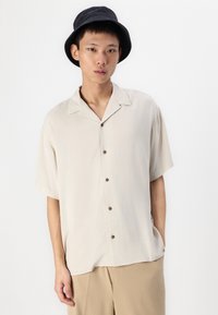 Short-sleeve beige button-up shirt with a camp collar, made of lightweight fabric, featuring brown buttons and relaxed fit design.