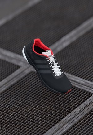 Chaussure de sport noire et rouge avec des lacets blancs flottant au-dessus d’un sol en grille métallique, montrant des bandes latérales et un détail de languette en mesh.