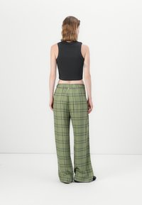 Woman shown from back wearing black sleeveless crop top and loose green plaid pants standing against white background.