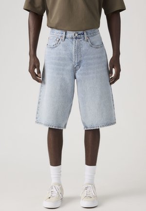 Mittellange hellblaue Jeansshorts, getragen von einer Person, kombiniert mit weißen Sportsocken, beigen Turnschuhen und einem olivgrünen Hemd.