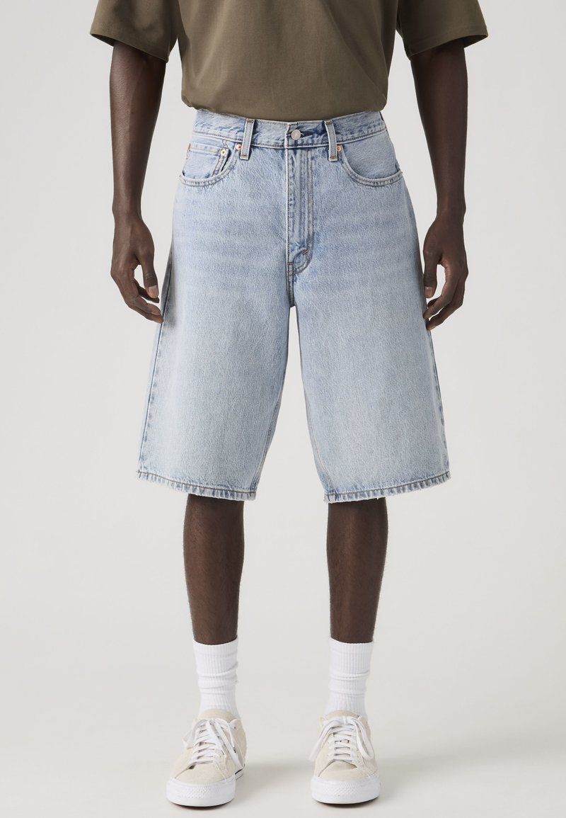 Mittellange hellblaue Jeansshorts, getragen von einer Person, kombiniert mit weißen Sportsocken, beigen Turnschuhen und einem olivgrünen Hemd.