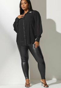 Camisa negra de botones con mangas abullonadas, cuello adornado, combinada con leggings negros de piel sintética y sandalias de tacón negras.