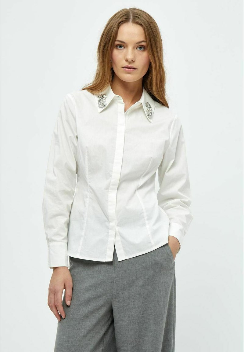 Camicia bianca in cotone con colletto decorativo in argento, maniche lunghe e design aderente, abbinata a pantaloni grigi.