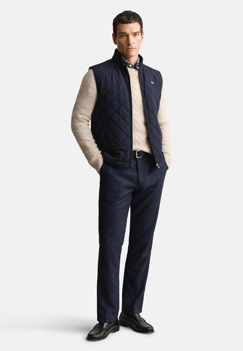 Chaleco acolchado azul marino sobre un suéter crema, combinado con pantalones azul oscuro y zapatos negros. Texturas suaves con detalle de cuello abotonado.