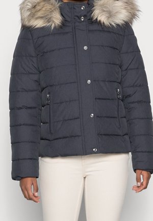 Navy blue padded winter jacket with fur-trimmed hood, snap buttons, and zippered side pockets worn with light beige pants.