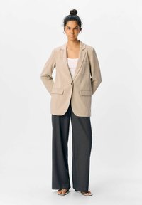 Kvinna står med händerna bakom ryggen, iklädd beige kavaj, vit topp, mörka vida byxor och sandaler med fyrkantig tå mot vit bakgrund.