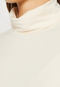 Crème coltrui gemaakt van zachte stof, met een nauwsluitende, opgerolde kraag en een gladde textuur, die dicht om de nek en schouders aansluit.