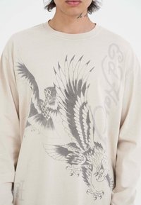 Beige Langarmhemd aus Baumwolle, mit aufwändigen grauen Adlergrafiken und auffälliger Schrift auf der Vorderseite.