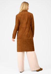 Frau von hinten trägt einen langen braunen Wildledermantel, cremefarbene weit geschnittene Hosen und braune Schuhe vor einem schlichten weißen Hintergrund.