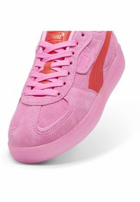 Sneaker in suede rosa con lacci e soletta coordinati. Presenta un logo PUMA rosso lucido e una striscia laterale. Suola in gomma arrotondata.