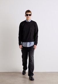 Sweatshirt preto com logo em relevo, sobre uma camisa azul clara. Jeans pretos e sapatos escuros, complementados por óculos de sol escuros.