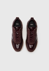 Un paio di sneakers BOSS lucide di un elegante bordeaux scuro, con un mix di suede e pelle, allacciatura frontale e suola bianca, su sfondo bianco.