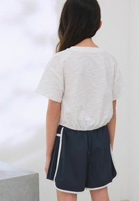 Haut court blanc texturé à manches courtes avec taille élastique, associé à un short bleu foncé avec bord blanc et poches. Vue arrière montrée.