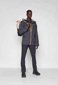 Tamsiai pilka vandeniui atspari striukė su geltonais užtrauktukais, pilka kapišonu ir derančiomis tamsiomis kelnėmis. Modelis nešioja smėlio spalvos ir juodą kuprinę.