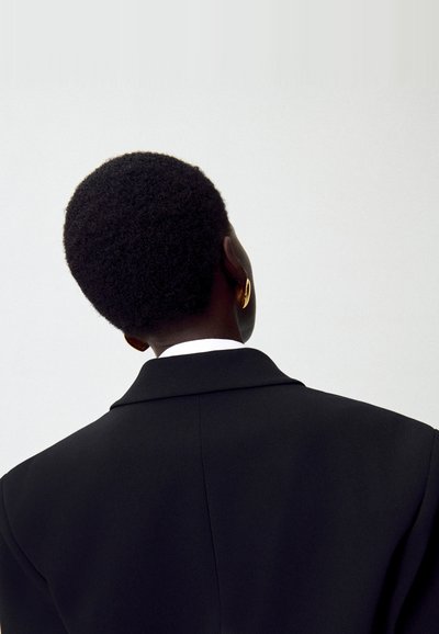 Blazer nero visto da dietro, con colletto strutturato, vestibilità su misura e una camicia bianca sotto. Orecchino a cerchio dorato visibile.